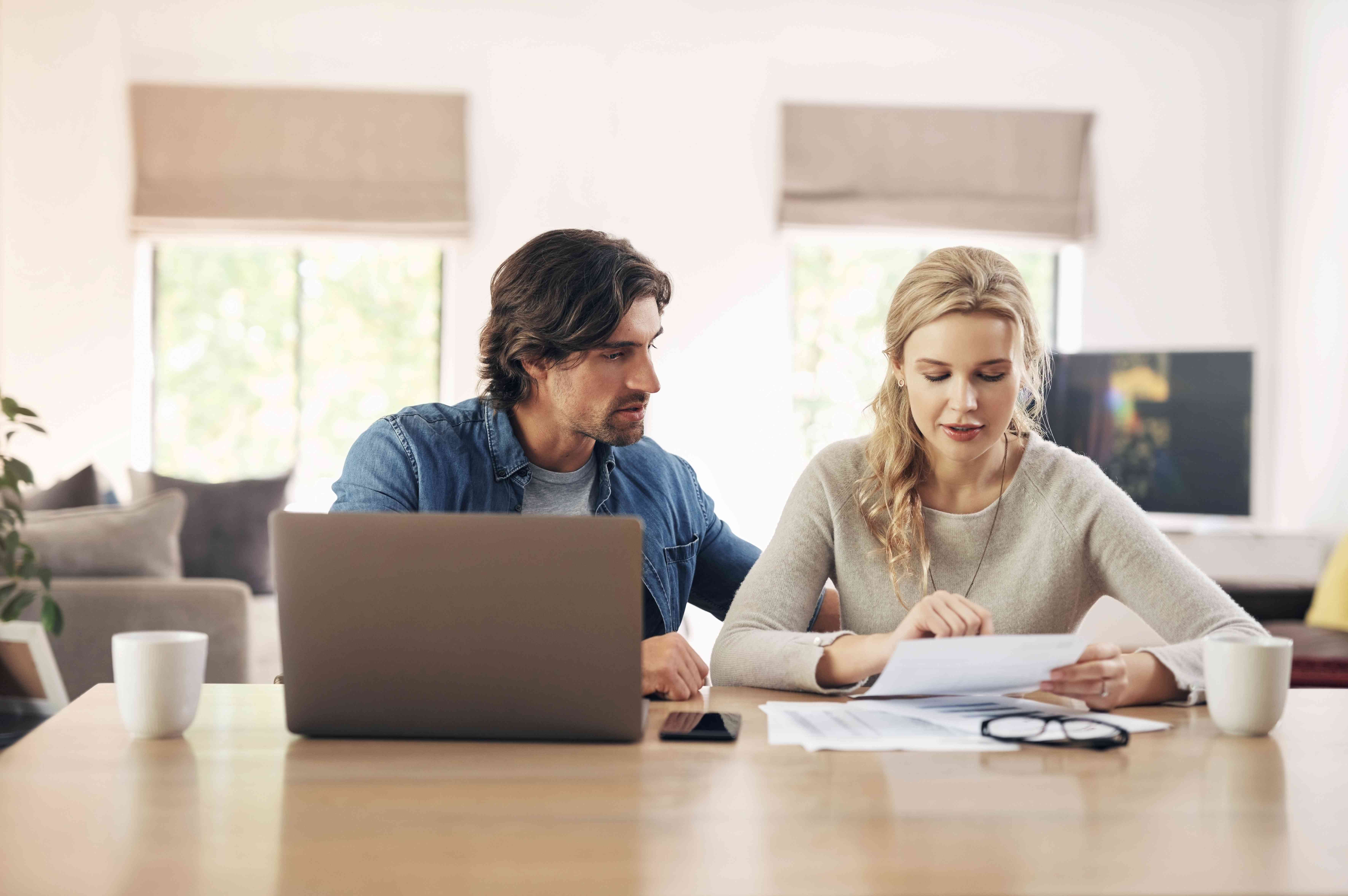 A man and a woman sit at a table with paperwork and a laptop, plus glasses and a coffee mug