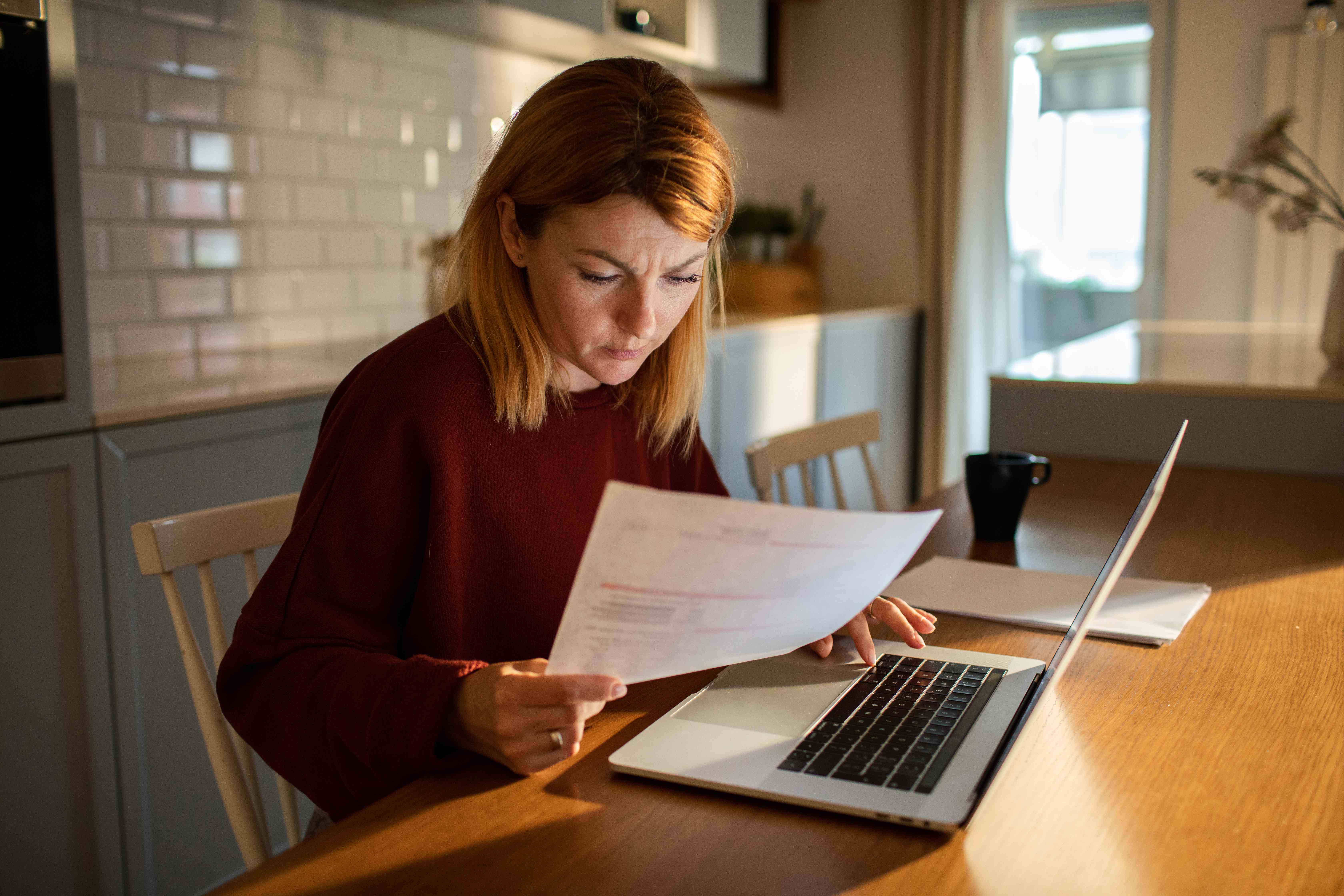 A young woman goes over her finances at a table, with paperwork and a laptop.